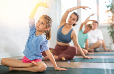 Pleasant little girl practicing half lotus pose of yoga raising one hand over head together with her family members