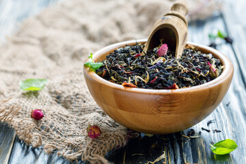 Black tea with flower petals and scoop in bowl on old wooden table, selective focus.