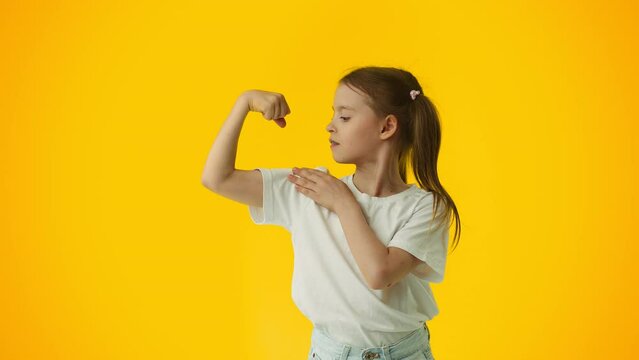 Young girl bending her arm, demonstrate her bicep and touch it on yellow background. Concept of proper child development, healthy nutrition and physical training