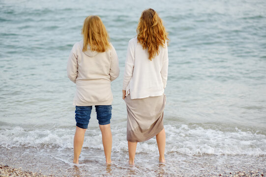 Red-haired Elderly Mother And Her Grown-up Daughter Are Walking Along The Seashore Together. Happy Meeting Of A Mother And Her Grown-up Child. Family Relations Between Adult Children And Parents