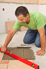 Man laying ceramic floor tiles - checking lines with a level, knealing on the concrete in a new...