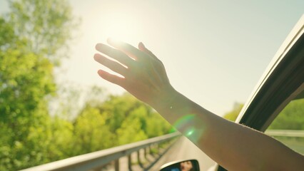 Free young girl waves her hand from car window, travels, catches sun glare with her fingers. Auto...