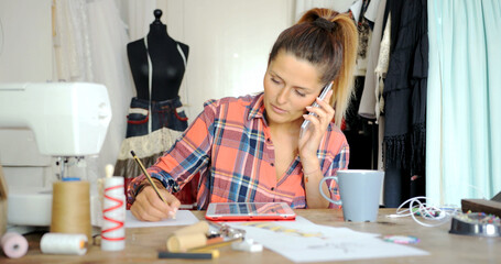 Fototapeta na wymiar Young female in casual clothing talking by phone while drawing sketch and using tablet in parlour.