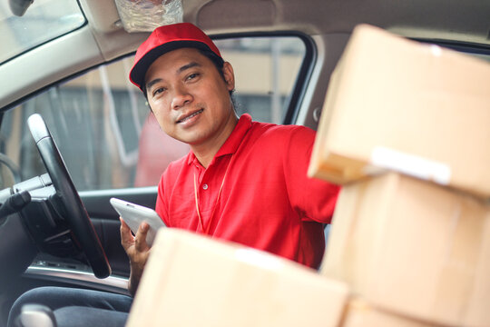 Asian deliveryman looking at camera and sitting in delivery van with pile of cardboard packages outside the warehouse