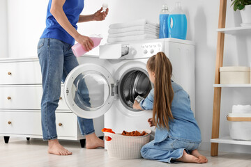 Mother pouring fabric softener and daughter putting dirty clothes into washing machine in bathroom, closeup
