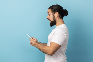 Side view of surprised man with beard wearing white T-shirt expressing shock while using cell phone, searching web, astonishing news. Indoor studio shot isolated on blue background.