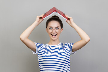 Portrait of woman wearing striped T-shirt and glasses holding opened book on head, looking at...