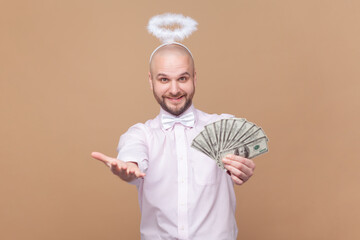 Portrait of satisfied rich bald bearded man with nimb over head, showing dollar banknotes, giving you salary, wearing light pink shirt and bow tie. Indoor studio shot isolated on brown background.