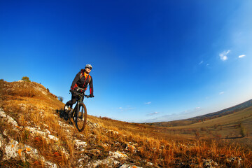 Fototapeta premium Wide angle view of a cyclist riding a bike on a nature trail in the mountains. people living a healthy lifestyle. Cyclist with helmet and gjasses. Beautiful landscape with hill and blue sky. Spring se