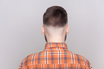 Back view portrait of dark haired bearded man with stylish hairstyle standing backwards to camera, looking far, wearing orange checkered shirt. Indoor studio shot isolated on gray background.