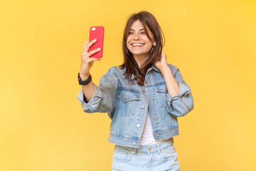 Portrait of charming pretty smiling woman blogger wearing denim jacket holding mobile phone in...