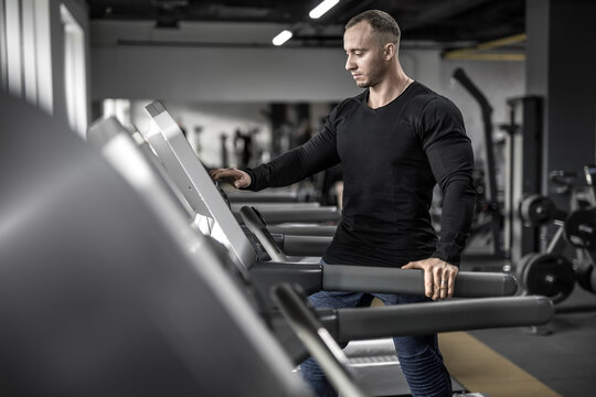 Brutal muscular man is preparing to run on the treadmill in the gym. Guy looks at the control panel. He wears a black sweatshirt and blue pants. Shoot from the side. Horizontal.