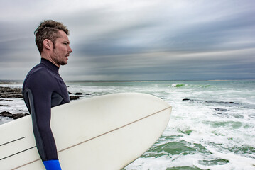 A side portrait of a surfer, with his surfboard under his arm as he views the ocean.