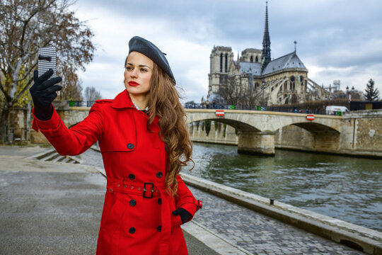 Bright in Paris. modern woman in red trench coat on embankment near Notre Dame de Paris in Paris, France with phone taking selfie