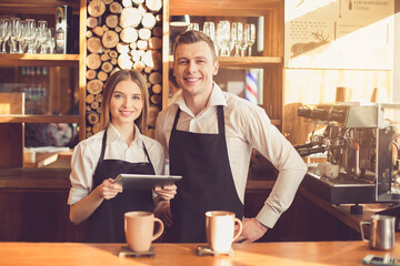 Professional barista. Young woman and man in aprons looking at camera, smiling and standing at bar counter. Woman using tablet computer