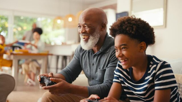 Grandfather and grandson sitting on sofa at home holding controllers playing video game together with multi-generation family in background - shot in slow motion