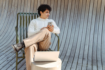 Smiling curly haired latin man holding mobile phone while resting at wooden armchair. Young happy...