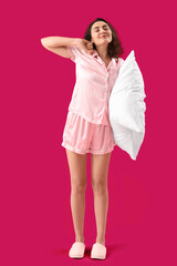 Young brunette woman in pajamas with pillow on pink background