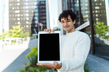 Happy smiling hispanic young businessman holding digital tablet, showing blank screen, standing in modern district. Successful man looking at camera. Business concept