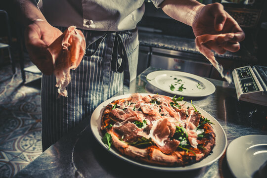 Food concept. Preparing traditional italian pizza. Chef in uniform decorate ready dish with green rucola herbs, ham, salami and prosciutto in interior of modern restaurant kitchen. Ready to eat.