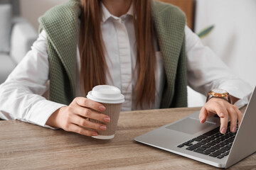 Woman using modern laptop and holding takeaway cup of coffee at table