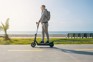 Young Caucasian man riding electric scooter along the sea coast near the beach at sunny summer day. Male driving e-scooter outdoors. Sustainable transport and travel