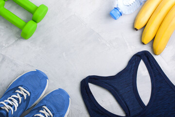 Flatlay sport composition with blue sneakers, t-shirt, green dumbbells, bananas and bottle of water on gray concrete background. Concept healthy lifestyle, sport, training in gym, diet. Horizontal ori