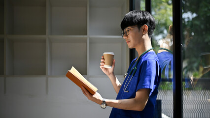 Rear view of a doctor leaning on the glass wall, sipping coffee, and reading a book in the office.