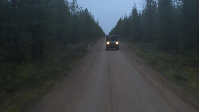 Side by side UTV races through dirt road in foggy pine tree forest with headlights on
