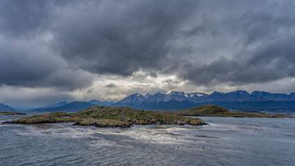 A rocky island with sparse vegetation in the sea strait. A picturesque snow-capped mountain range against a cloudy sky. Argentina. Tierra del Fuego Archipelago. Beagle Channel