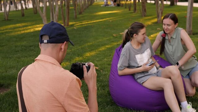 A father photographs his family in a crowded city park in summer, a mother and daughter sit in soft chairs on the grass and look at the phone.