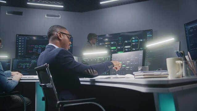 Multiracial IT technical specialists sit in front of computers with data server in monitoring office. Software engineer works with live analysis charts and blockchain network on big digital screens.