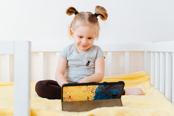 Smiling little caucasian girl watching cartoons on tablet, cute child using technology while sitting on bed in children's room