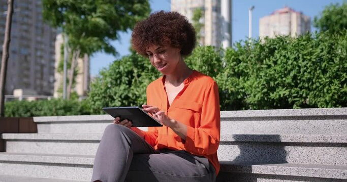 Happy young African American woman using digital tablet pad sufring internet inspired with new ideas sitting outdoors