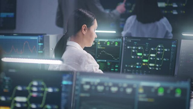 Female software engineer works on computer with data server and blockchain network database in modern monitoring control room. Team of IT technical specialist and big digital screens on background.