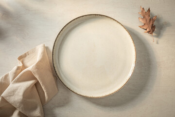 Empty ceramic plate, linen napkin and autumn leaf on stone background in warm monochrome