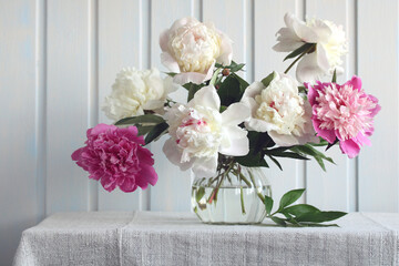 Still life with a bouquet of peonies in a glass vase on a table against a white wooden wall.