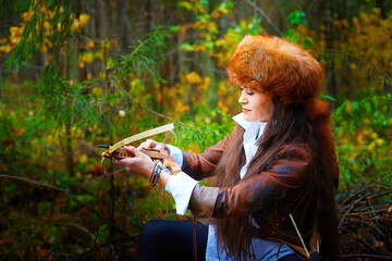 Girl in a leather jacket, a big red fox fur hat and with a crossbow in the forest in autumn. A...
