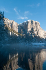 Fototapeta na wymiar The rocky surface of the Sierra Nevada mountains, reflecting early morning sunlight, being reflected in Merced river, on an early winter, snow-covered landscape in Yosemite national park.