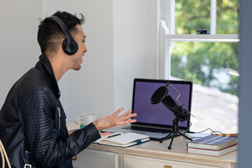 Happy biracial transgender man in headphones, using laptop, talking to microphone while podcasting