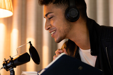 Happy biracial transgender man in headphones, making notes and talking to microphone, podcasting