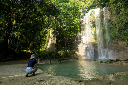 An asian tourist in a blue top and jorts taking a picture of a waterfall. At Kawasan falls in Balilihan, Bohol.