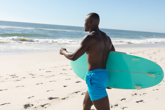 Fit african american man carrying surfboard walking on sunny beach to sea
