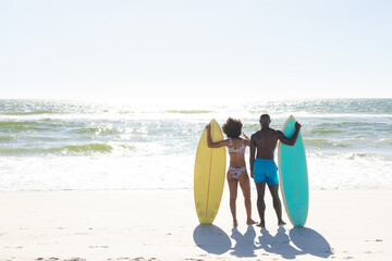 Rear view of african american couple with surfboards standing on sunny beach facing sea, copy space