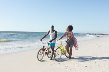 Obraz na płótnie Canvas Happy african american couple walking with bikes on sunny beach by sea