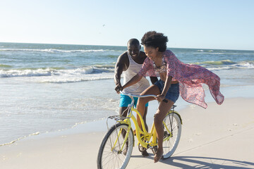 Happy african american couple having fun walking and riding bike on sunny beach by sea, copy space