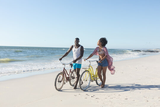Happy african american couple walking with bikes on sunny beach by sea