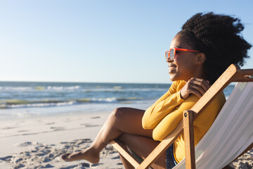 Happy african american woman in sunglasses sitting in deckchair smiling on sunny beach by sea