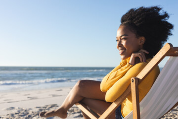 Happy african american woman sitting in deckchair smiling on sunny beach by sea