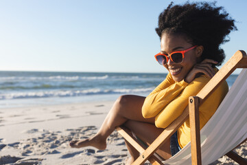 Portrait of african american woman in sunglasses sitting in deckchair smiling on sunny beach by sea